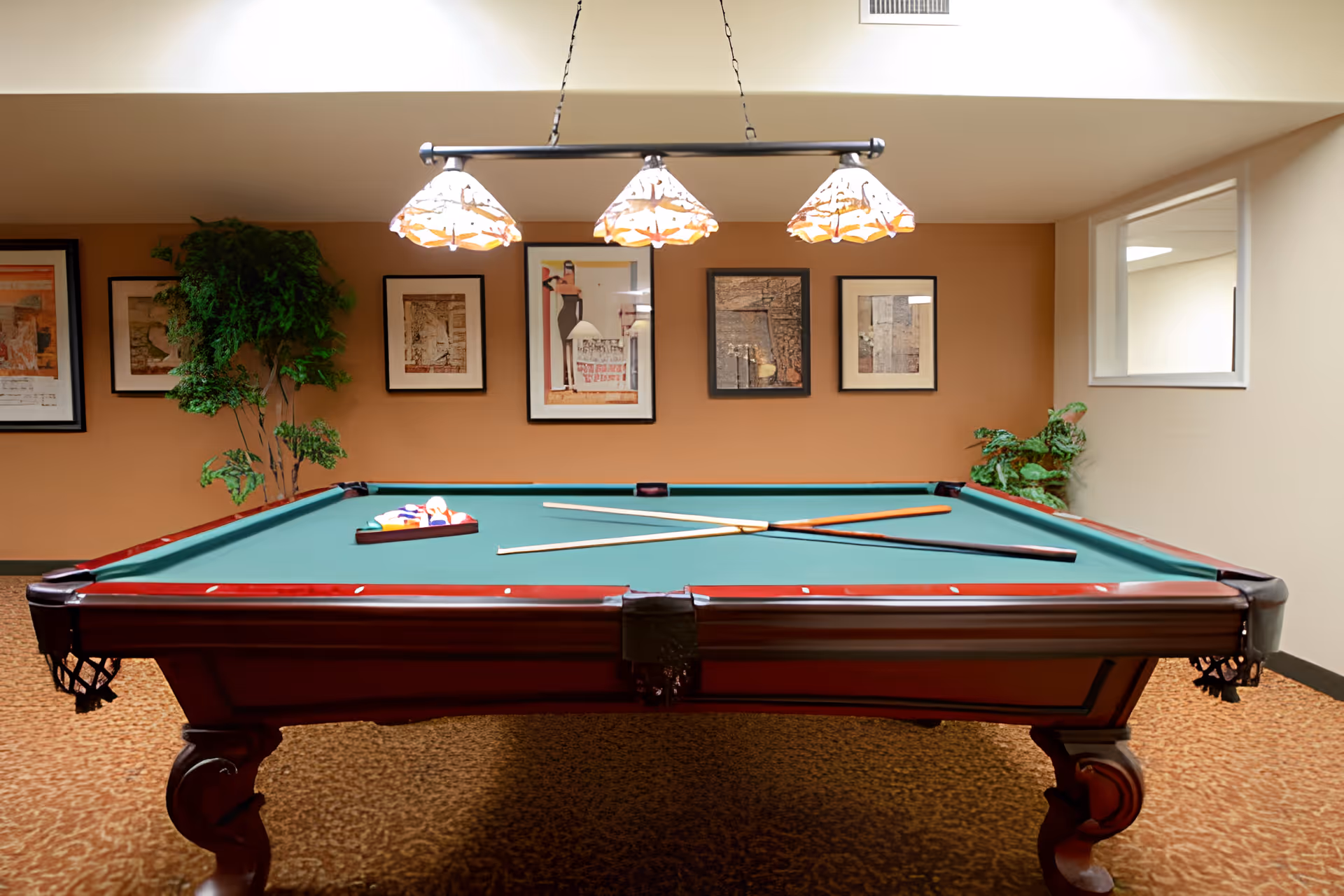 A pool table with green felt and wooden frame is centered in a room with beige walls and carpeted floor. Three pool cues and a triangle rack with billiard balls are placed on the table. Above the table, a light fixture with three decorative lampshades hangs from the ceiling. The back wall features five framed artworks and two potted plants are positioned on either side of the wall. A window is visible on the right wall.