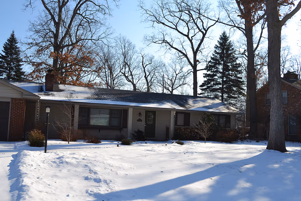 Single-story house with a snow-covered yard and leafless trees in the background under a clear blue sky.