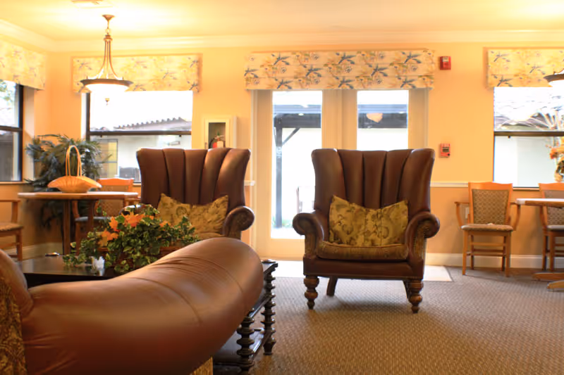 A warm, well-lit lounge with two wingback leather armchairs, a leather sofa, tables, and floral decorations in front of glass doors.
