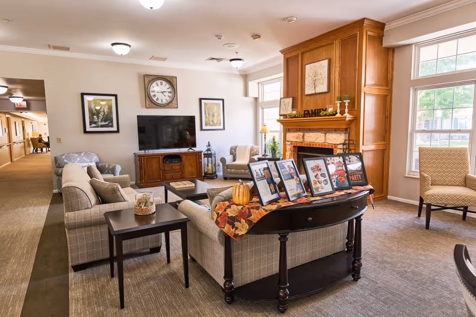 A cozy living room area in a senior living facility with beige and plaid upholstered sofas and armchairs arranged around a dark wood coffee table and side tables. A large flat-screen TV is mounted on a wooden cabinet against the wall, flanked by framed botanical prints and a large clock above. A wooden fireplace with a mantle decorated with candles, greenery, and framed art is on the right side. A console table behind the sofa displays framed informational signs and a small pumpkin on a fall-themed table runner. Large windows let in natural light, and a hallway is visible to the left.