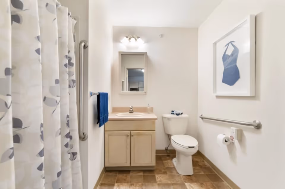 A clean and simple bathroom featuring a shower with a patterned curtain on the left, a vanity with a sink and mirror in the center, and a toilet on the right. The walls are white, and there is a blue towel hanging on a rack next to the vanity. A framed picture of a blue swimsuit hangs on the wall above a grab bar next to the toilet. The floor has brown tiles.