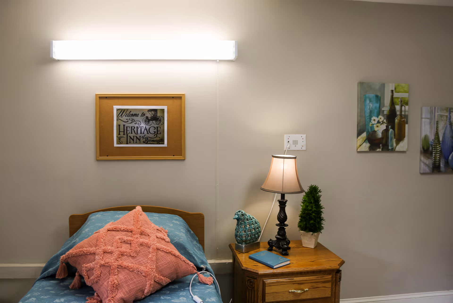A single bed with a blue bedspread and a large pink textured pillow is positioned against a beige wall. Above the bed, there is a framed sign that reads 'Welcome to Heritage Inn of Barnesville.' Next to the bed is a wooden nightstand with a small lamp, a decorative blue bird figurine, a small potted plant, and a closed blue book. Two paintings featuring bottles and flowers hang on the wall to the right.