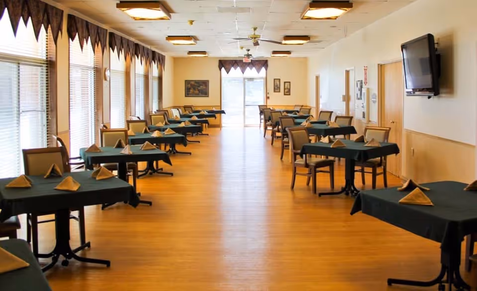 Spacious dining room with rows of tables covered in green tablecloths and folded napkins, chairs, and large windows along one side.