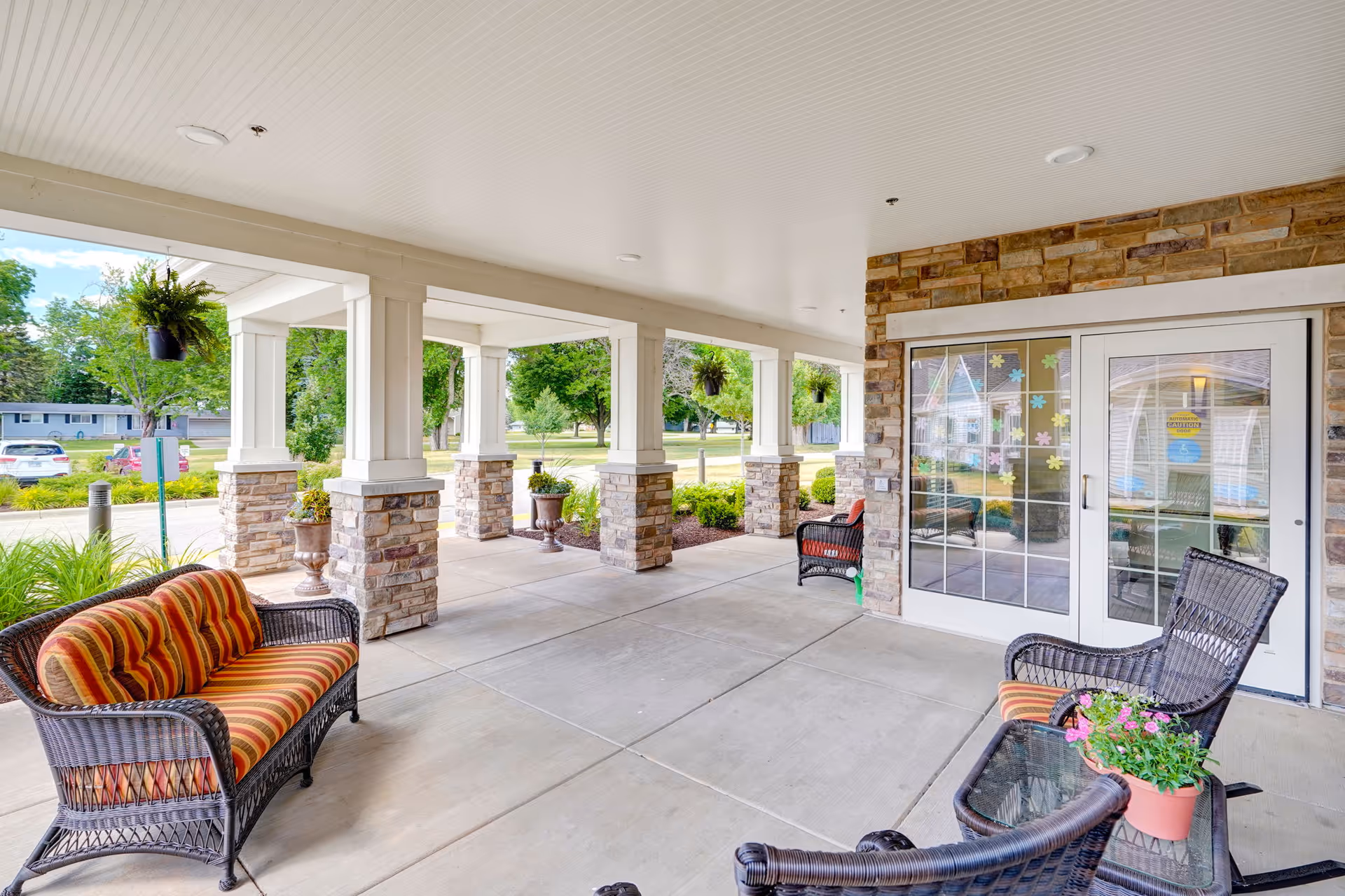 Covered outdoor patio area with wicker furniture including a striped cushioned loveseat, chairs, and a glass-top table with a potted plant. Stone pillars support the roof, and hanging plants are suspended from the ceiling. Glass double doors lead inside, decorated with colorful flower stickers. Green trees and a parking area are visible in the background.