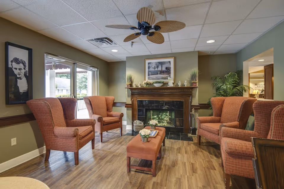 A cozy living room area with four red upholstered armchairs arranged around a wooden fireplace. The fireplace mantel is decorated with plants, figurines, and a framed picture above it. A ceiling fan with wicker blades hangs from the ceiling. There is a small ottoman with flowers and books in the center of the seating area. Large windows on the left side let in natural light, and a framed black and white portrait hangs on the wall near the windows.