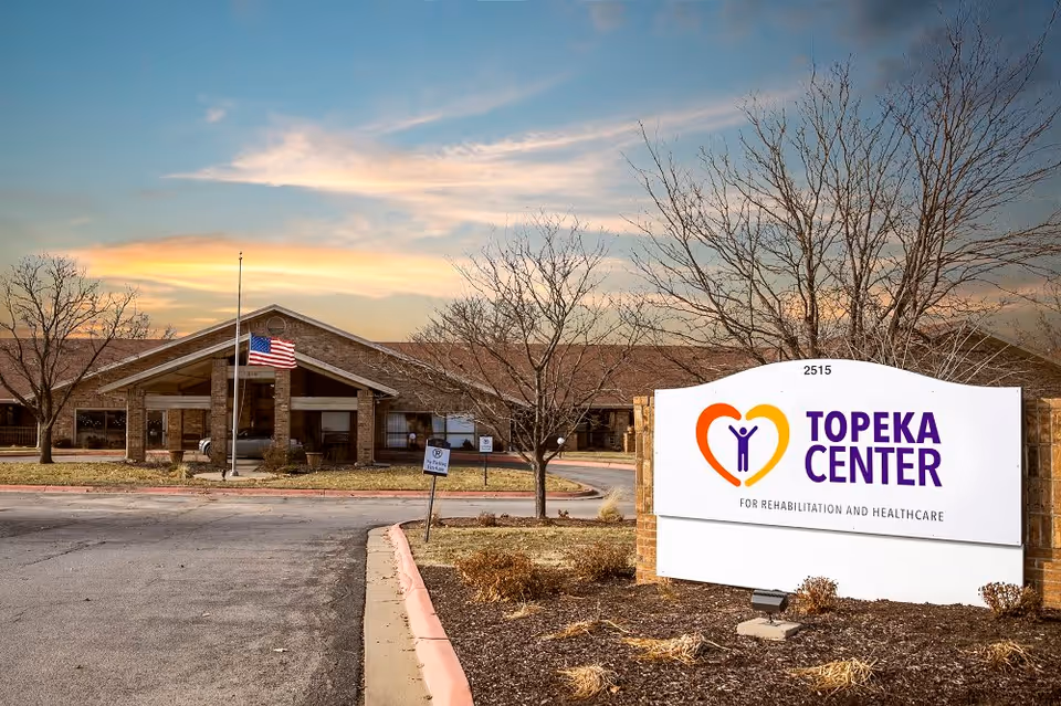 Exterior view of Topeka Center for Rehabilitation and Healthcare building during sunset with an American flag on a flagpole and leafless trees in the foreground. A large sign with the facility's name and logo is prominently displayed near the entrance.