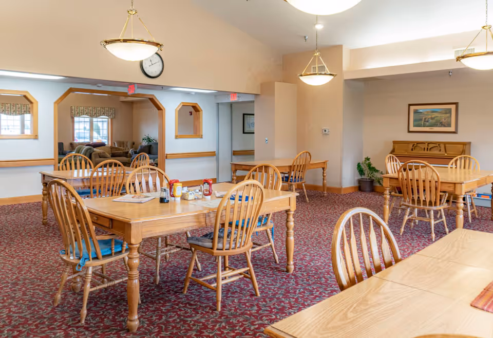 Dining room with wooden tables and chairs, pendant lights, and patterned carpet in a senior living facility.