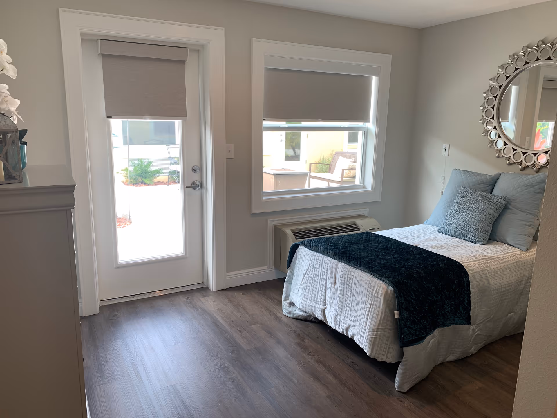 A small, neatly arranged bedroom with a single bed covered in white and dark blue bedding. The room has a window and a glass door with gray roller blinds, allowing natural light to enter. There is a decorative round mirror on the wall above the bed and a wooden floor.