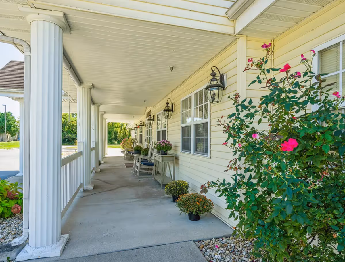 Covered porch area of a building with white columns, beige siding, and several potted plants and flowers. There are outdoor chairs and small tables along the wall under hanging lantern-style lights. A bush with pink flowers is visible on the right side.