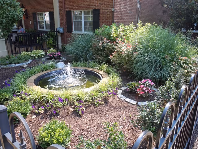 A landscaped garden area with a small circular water fountain in the center, surrounded by various green plants and flowers. The garden is enclosed by a black metal fence, and a brick building with windows and a porch is visible in the background.