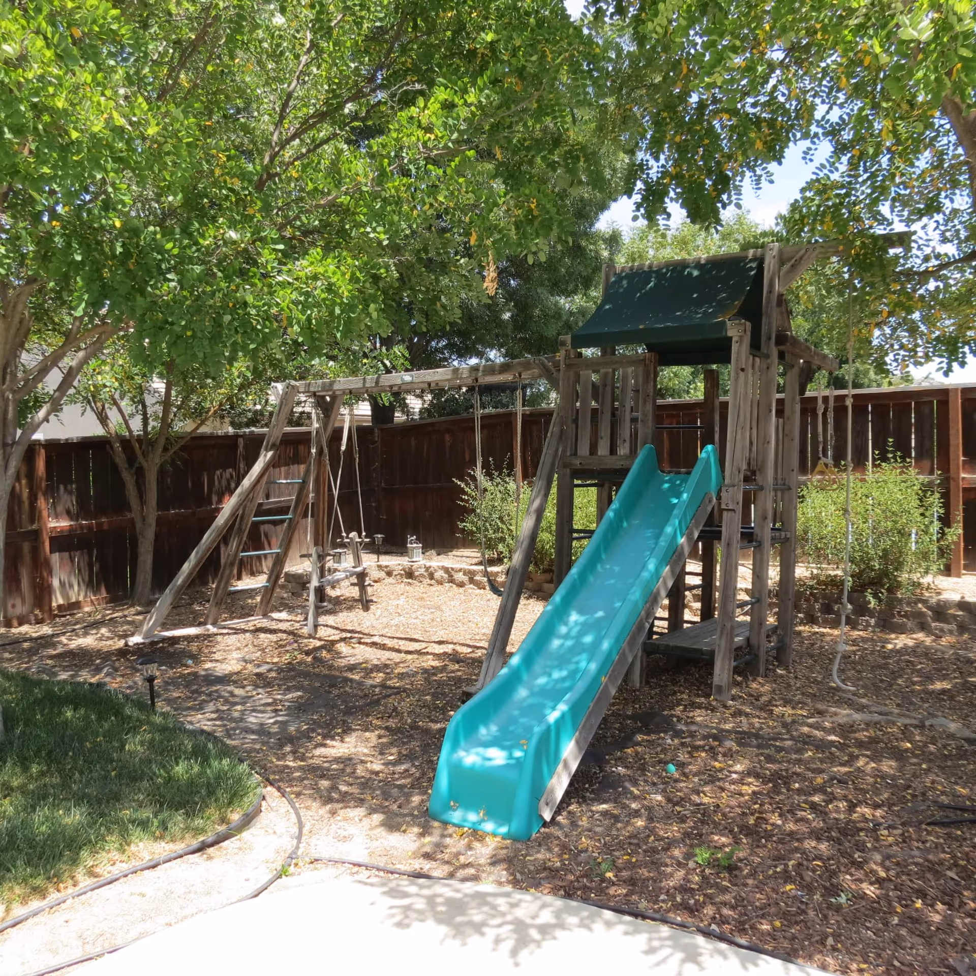Outdoor playground area with a wooden play structure featuring a green slide, swings, and a small climbing ladder, surrounded by trees and a wooden fence.
