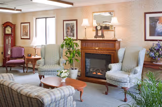 A cozy living room with two light blue patterned armchairs, a striped sofa, a wooden coffee table with a small plant, a wooden fireplace with a clock and lamp on the mantel, framed artwork on the walls, a grandfather clock, and several potted plants near the windows.