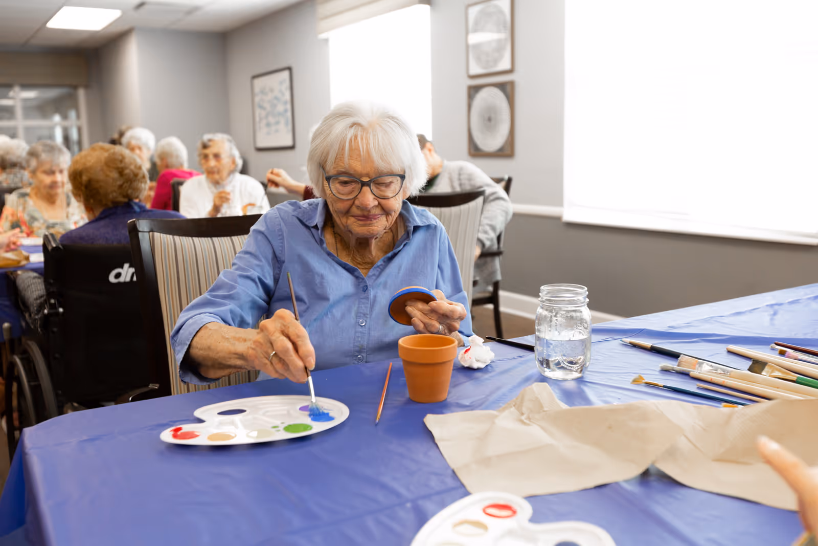 An elderly woman with white hair and glasses is painting a small terracotta pot blue at a table covered with a blue plastic tablecloth. She is holding a paintbrush in one hand and the pot lid in the other. There are paint palettes, brushes, a jar of water, and paper towels on the table. In the background, other elderly people are seated and engaged in activities in a well-lit room with gray walls and framed artwork.