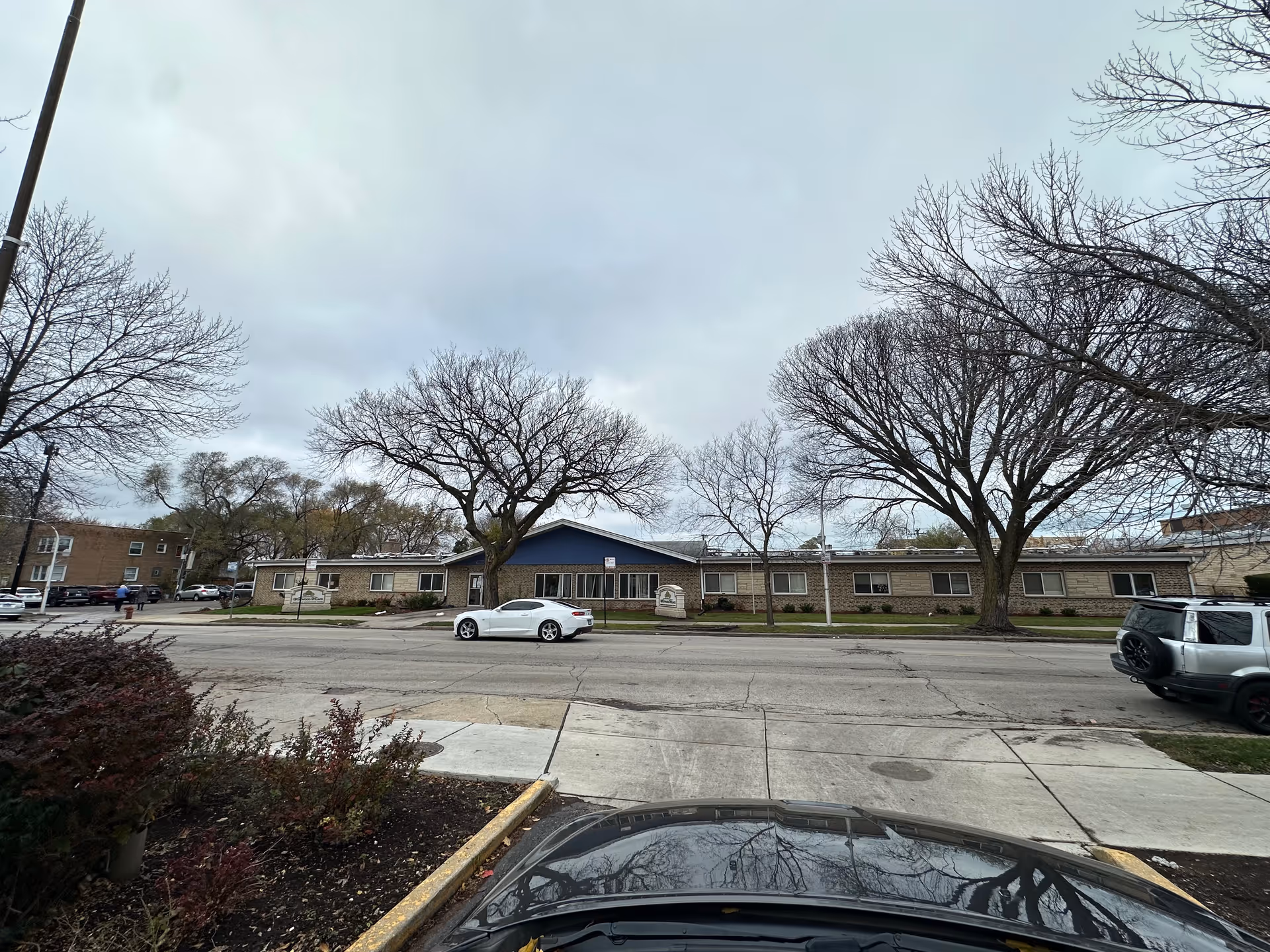 Exterior view of Westwood Village Nursing and Rehabilitation Center, a single-story brick building with a blue gable roof section in the center. There are leafless trees in front of the building and several cars parked along the street and in the parking lot. The sky is overcast.