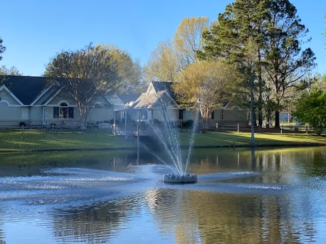 A serene pond with a water fountain spraying water upwards in the center, surrounded by green grass, trees, and residential buildings in the background under a clear blue sky.