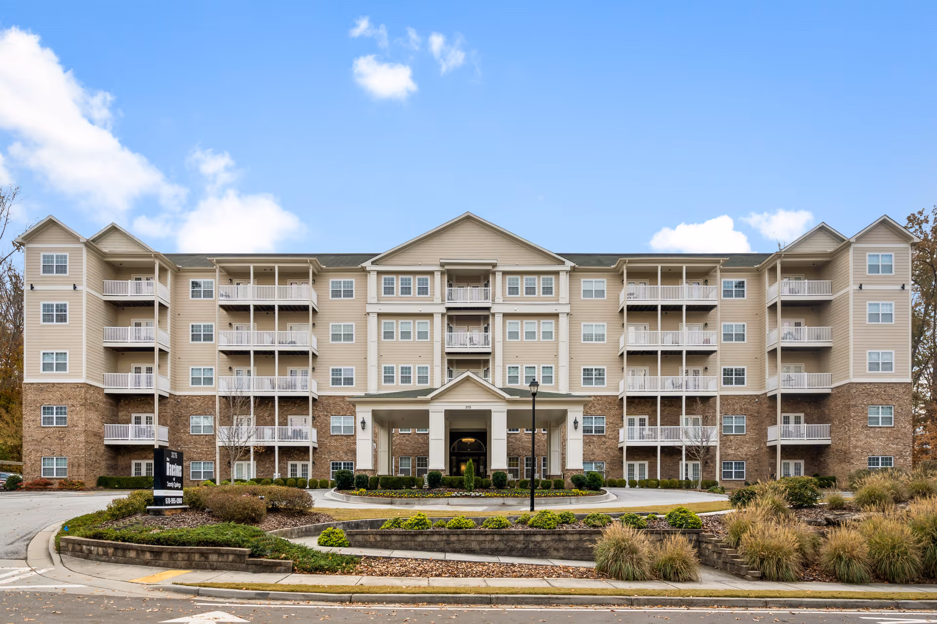 Front exterior view of a four-story senior living facility building with beige siding and stone accents, multiple balconies, a covered entrance, landscaped greenery, and a clear blue sky with some clouds.