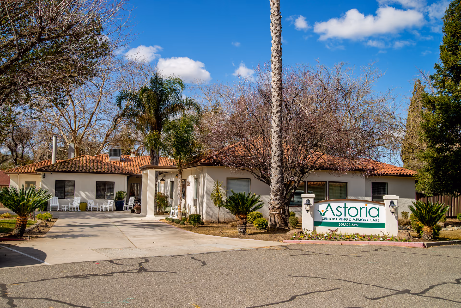 Exterior view of Astoria Senior Living facility with a single-story building featuring a red tile roof, surrounded by palm trees and other greenery under a blue sky with some clouds. A sign in front reads 'Astoria Senior Living & Memory Care' with a phone number.