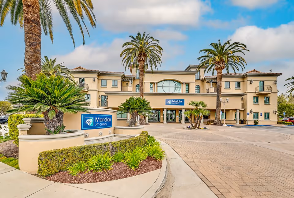 Exterior view of Meridian at Chino senior living facility showing a large beige building with multiple windows and palm trees in front. There is a curved driveway and landscaped greenery with signs displaying the facility name.