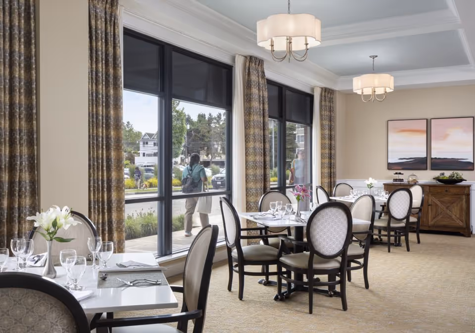 A bright dining room with several tables set with glassware, napkins, and silverware. The room features large windows with patterned curtains, allowing natural light to fill the space. There are upholstered chairs around the tables, two ceiling light fixtures, and a wooden sideboard against the far wall with decorative items and two framed landscape paintings above it. Outside the windows, people and houses are visible.