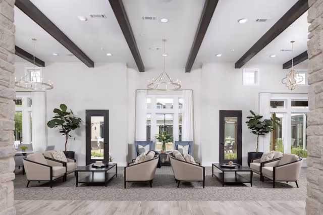 A spacious and bright living room area with high ceilings featuring dark wooden beams. The room is furnished with multiple beige armchairs arranged around black coffee tables on a patterned carpet. Large windows with white curtains allow natural light to fill the space, and there are two tall green plants in black pots on either side of the room. Modern chandeliers hang from the ceiling, and stone pillars frame the view.