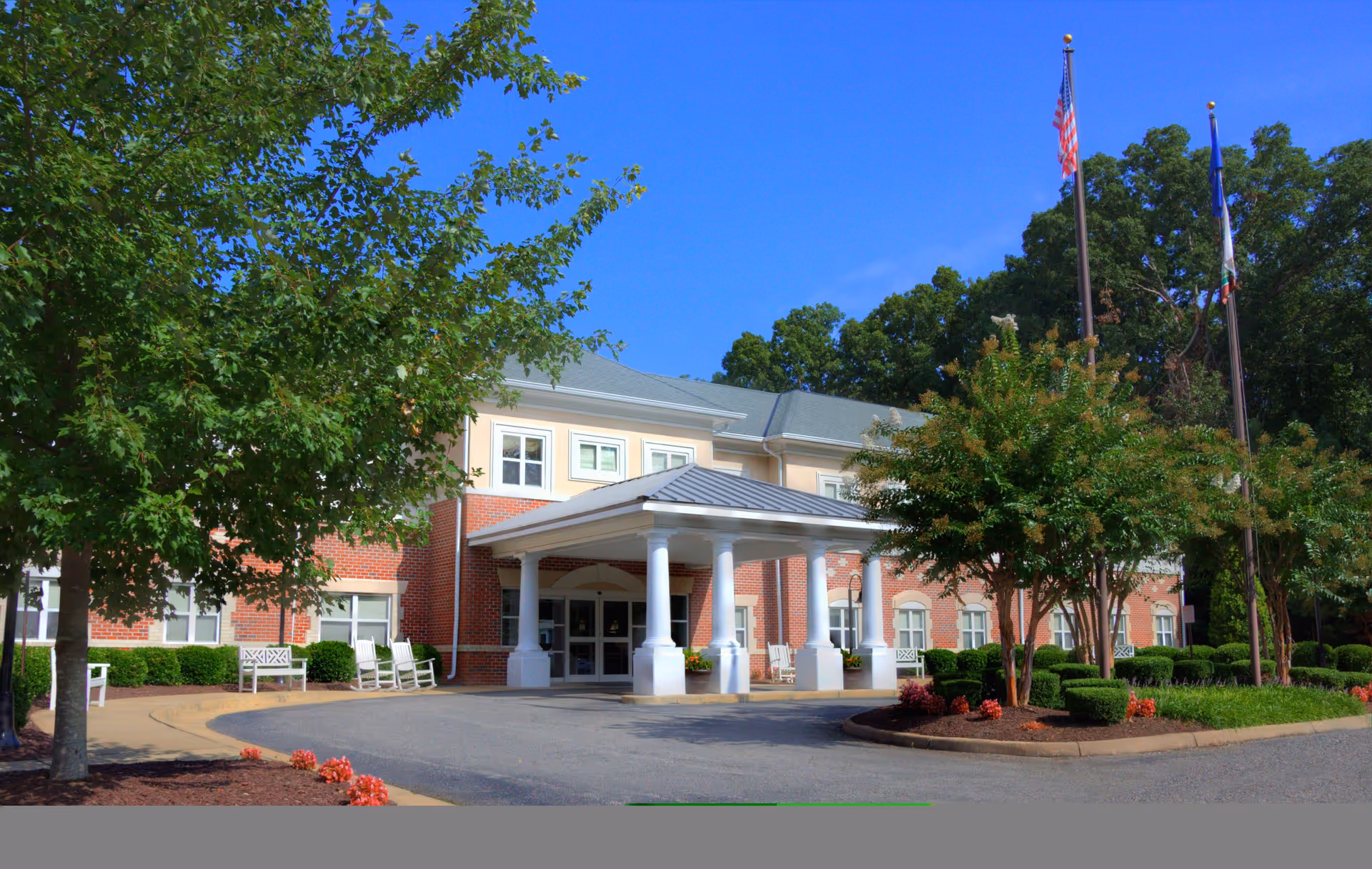 Front exterior view of a senior living facility building with a covered entrance supported by white columns, surrounded by trees, shrubs, and a clear blue sky. Two flagpoles with flags are visible near the entrance.