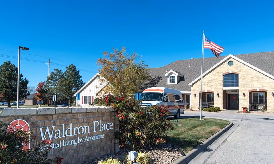 Front entrance of Waldron Place senior living facility with a stone sign, flagpole, shuttle van, and landscaping under a clear blue sky.