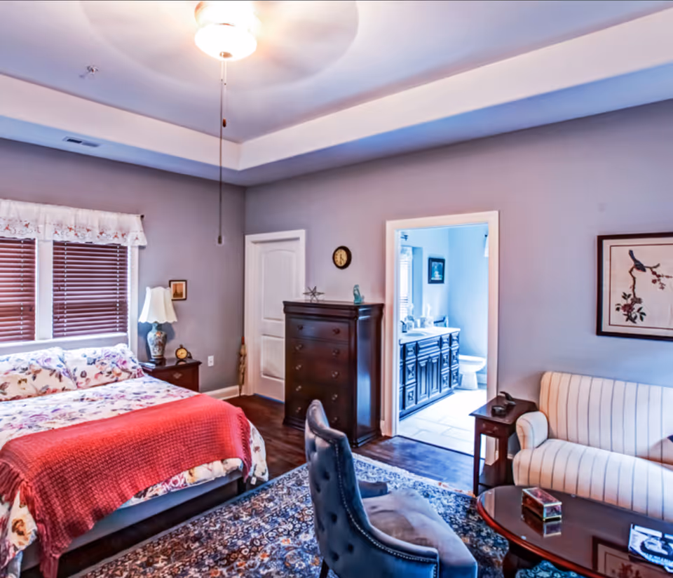 A cozy bedroom with a floral bedspread and red blanket on the bed, a wooden dresser, a nightstand with a lamp, and a ceiling fan. The room has gray walls and dark wood flooring. There is a doorway leading to a bathroom with a dark vanity and a toilet visible. A striped armchair and a wooden coffee table with decorative items are also present in the room.