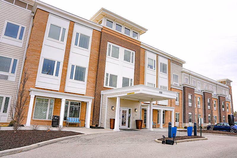 Exterior view of a three-story senior living facility building with a covered entrance, multiple windows, and a driveway in front. The building features a combination of brick and light-colored siding with some landscaping and parked cars visible.