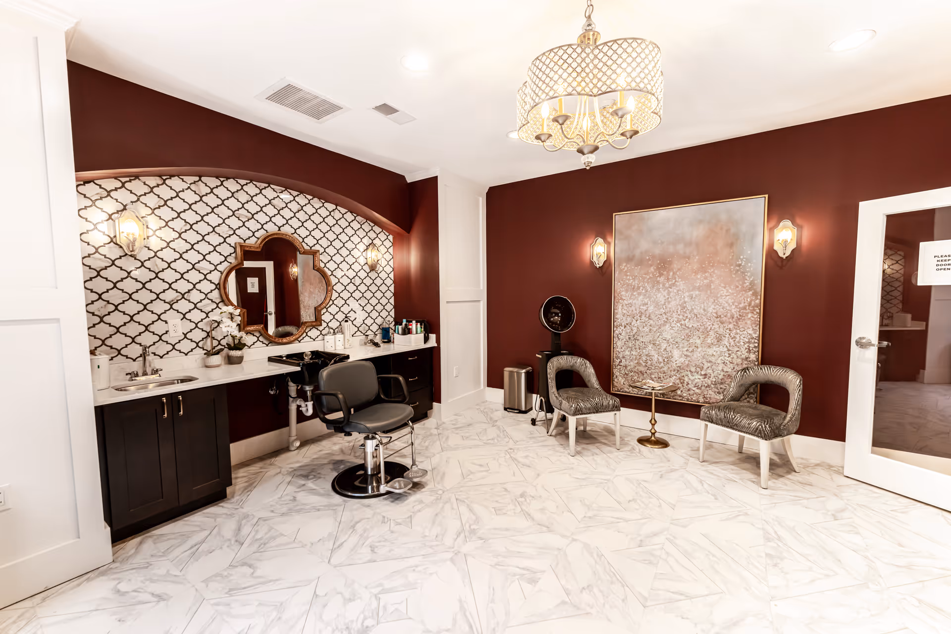 A stylish salon room with a black salon chair in front of a counter with a sink and mirror. The walls are painted dark red with a patterned tile backsplash behind the counter. Two decorative chairs and a large abstract painting are on the opposite wall. The floor is white marble tile, and a chandelier hangs from the ceiling.