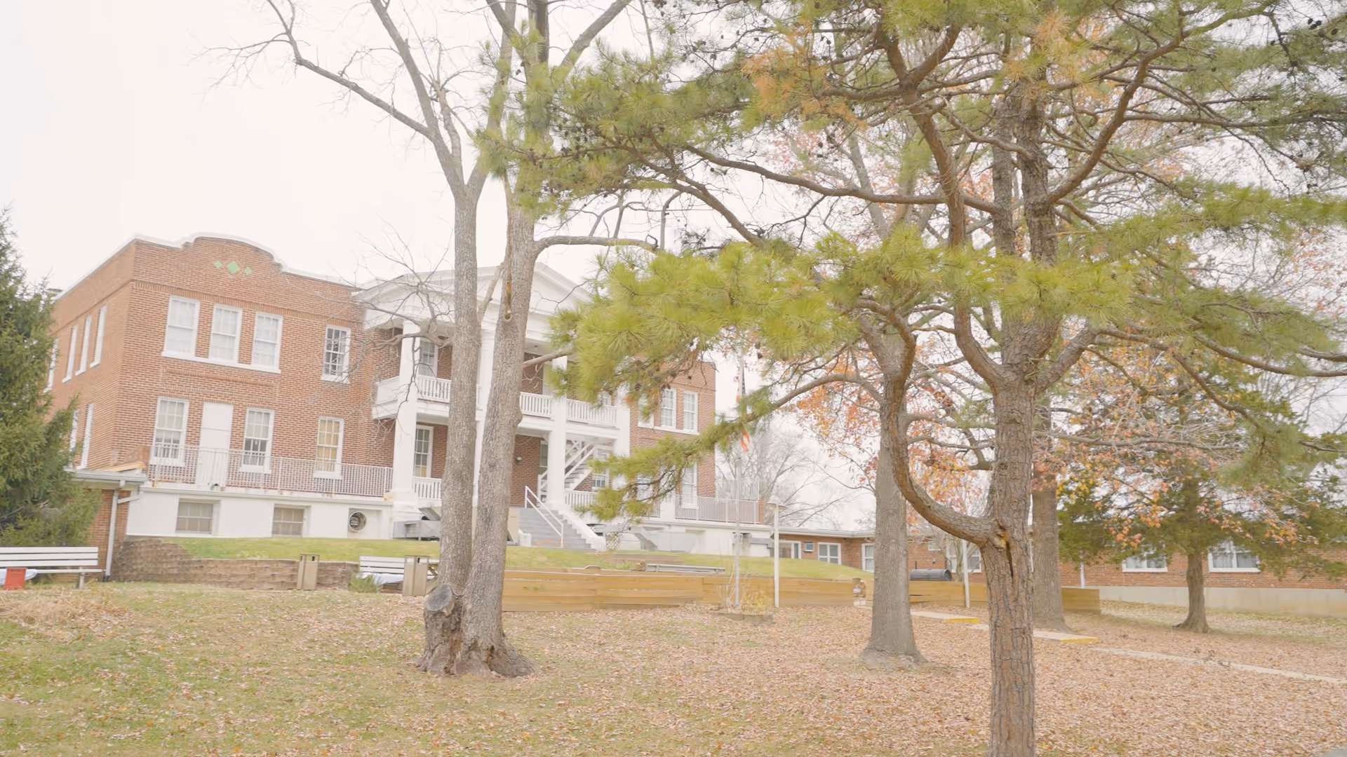 Exterior view of a brick building with white columns and multiple windows, surrounded by trees with sparse leaves and a grassy area covered with fallen leaves in the foreground.