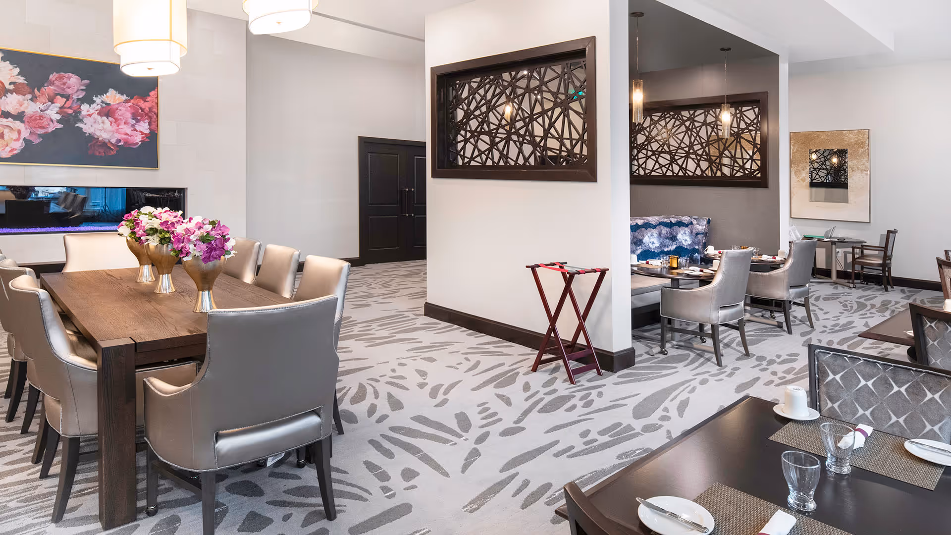 Bright, modern communal dining room with multiple tables, upholstered chairs, floral centerpieces, patterned carpet, and decorative wall art.