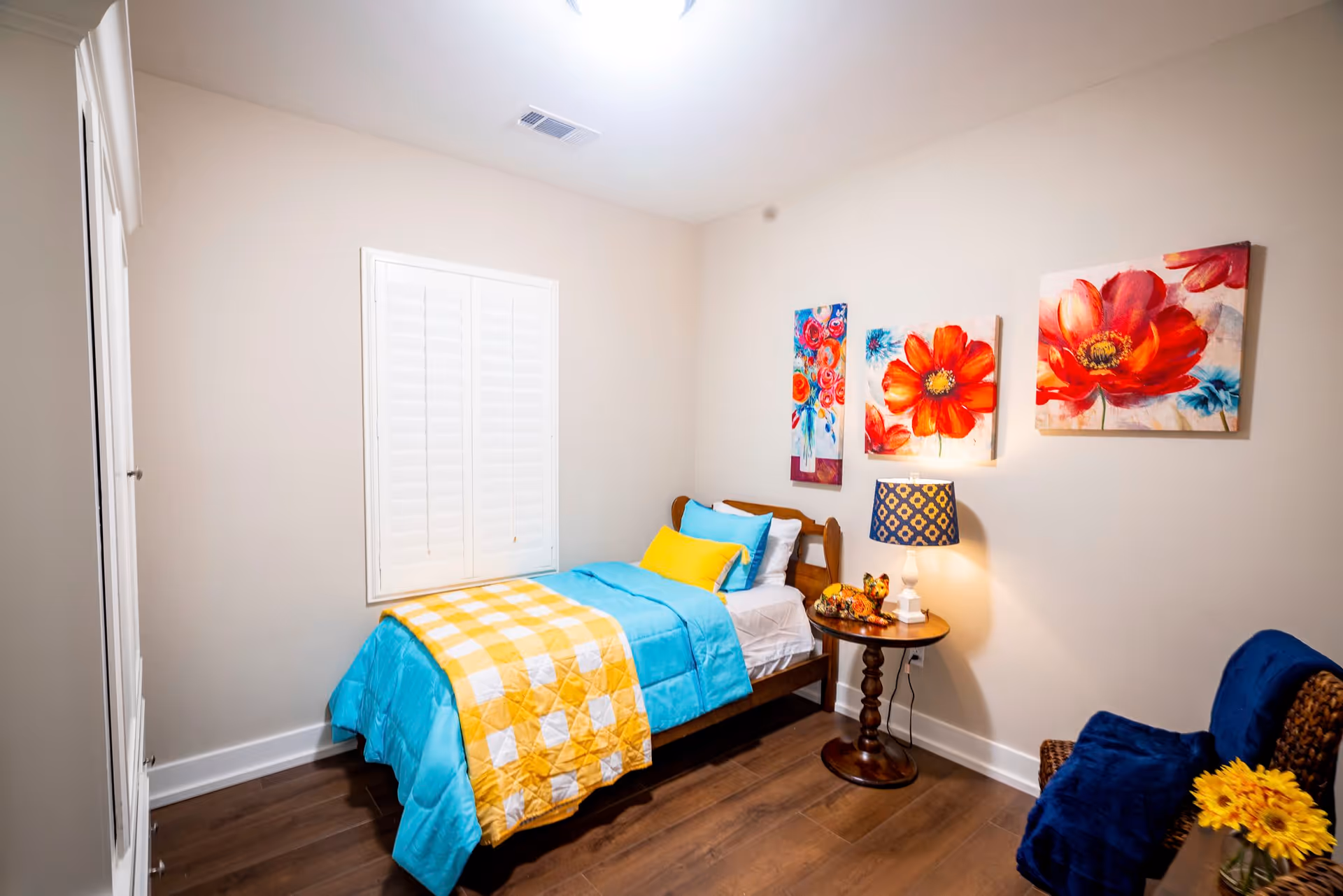 A small bedroom with a single bed dressed in bright blue and yellow bedding. The room has wooden flooring, a window with white shutters, and three colorful floral paintings on the wall above a round wooden side table with a lamp and decorative items. There is also a wicker chair with a blue blanket and a small bouquet of yellow flowers.
