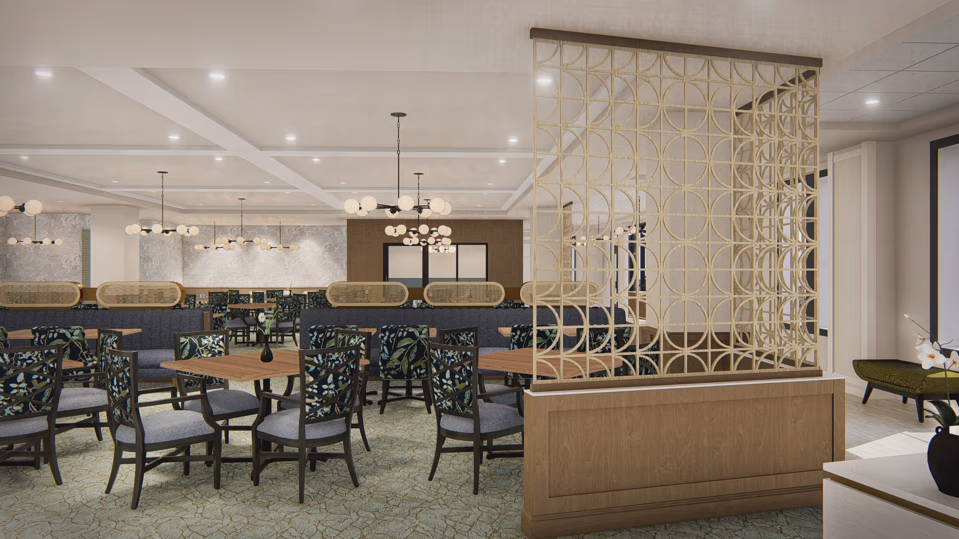 A spacious dining room with multiple wooden tables and chairs featuring floral upholstery. The room is decorated with modern chandeliers and has a decorative wooden and metal partition dividing the space. The walls are light-colored with subtle patterns, and the floor is covered with a patterned carpet.
