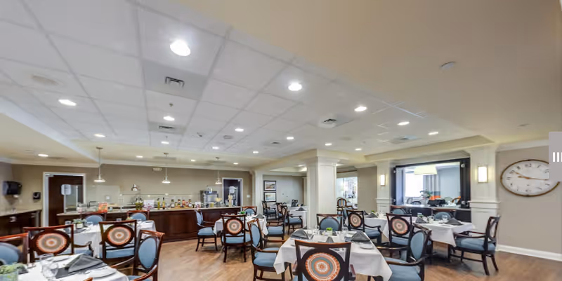 A spacious dining room with multiple tables covered in white tablecloths, each set with plates, glasses, and napkins. The chairs have decorative backs with a circular pattern. The room has a wooden floor, a drop ceiling with recessed lighting, and a buffet area with various food and drink items. A large clock is mounted on the wall to the right.
