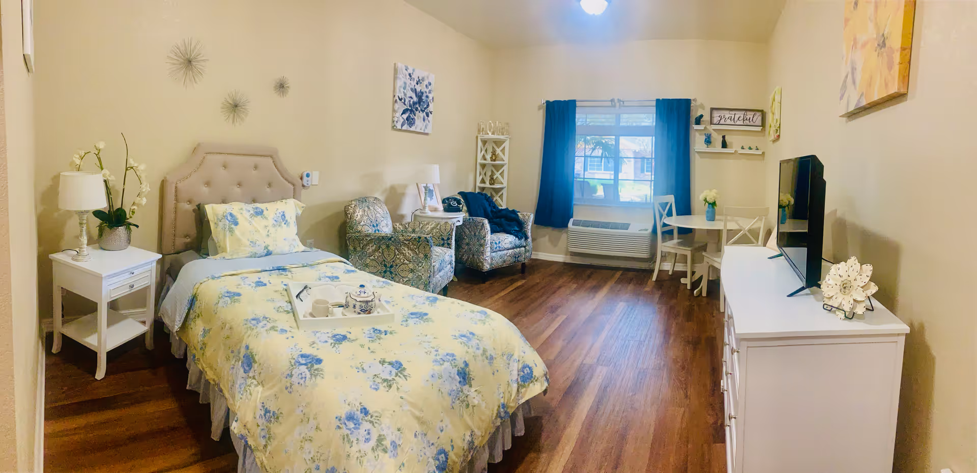 A cozy senior living bedroom with a single bed covered in yellow and blue floral bedding. Next to the bed is a white nightstand with a lamp and a plant. There are two patterned armchairs near a window with blue curtains. A small round table with two chairs is placed near the window. Across from the bed is a white dresser with a flat-screen TV on top. The room has wooden flooring and light-colored walls decorated with artwork and wall decor.