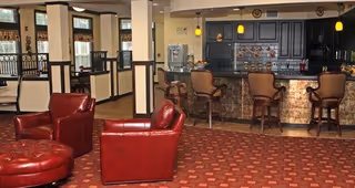 Interior view of a lounge area with red leather armchairs and a matching ottoman on a patterned carpet. In the background, there is a bar counter with four high chairs, pendant lights hanging above, and a coffee or beverage station. The space has large windows with curtains and wooden dividers separating different seating areas.