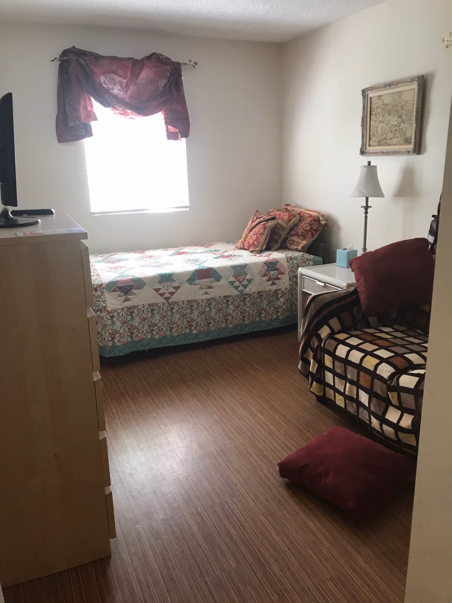 A small bedroom with a single bed covered in a colorful quilt and multiple pillows. Next to the bed is a white nightstand with a lamp and a box of tissues. A patterned armchair with a red cushion is positioned near the nightstand, and another red cushion is on the floor. A wooden dresser with a TV on top is on the left side of the room. The window has a dark red valance curtain, and a framed picture hangs on the wall above the lamp.