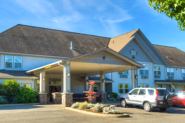 Exterior view of a senior living facility building with a covered entrance and several parked cars in front. The building has light-colored siding, multiple windows, and a sloped roof under a clear blue sky.