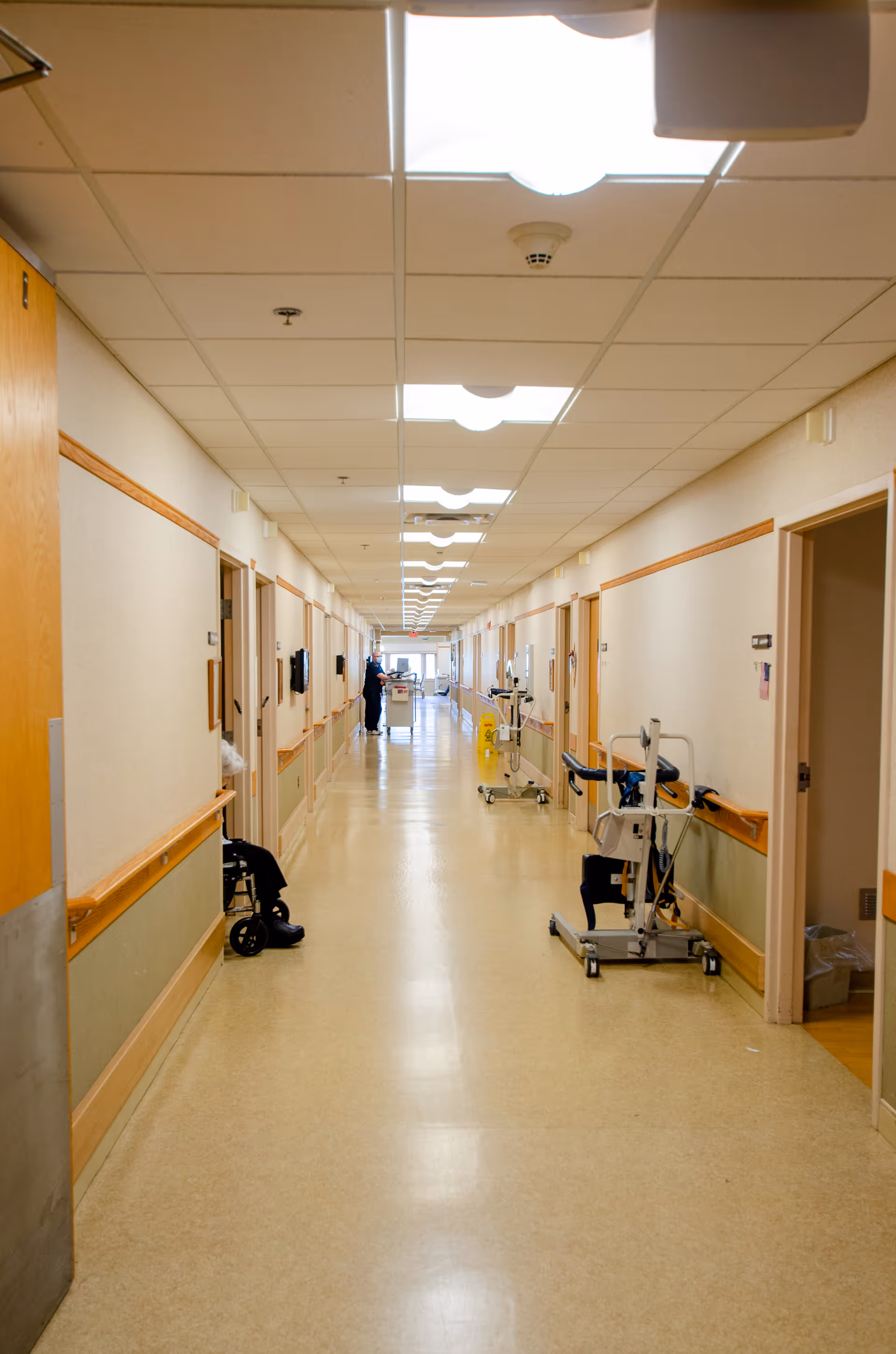 Long, well-lit hallway in a rehabilitation and retirement community with beige walls and floor, handrails along both sides, several open doors, mobility aids such as wheelchairs and walkers placed along the walls, and a staff member visible in the distance.