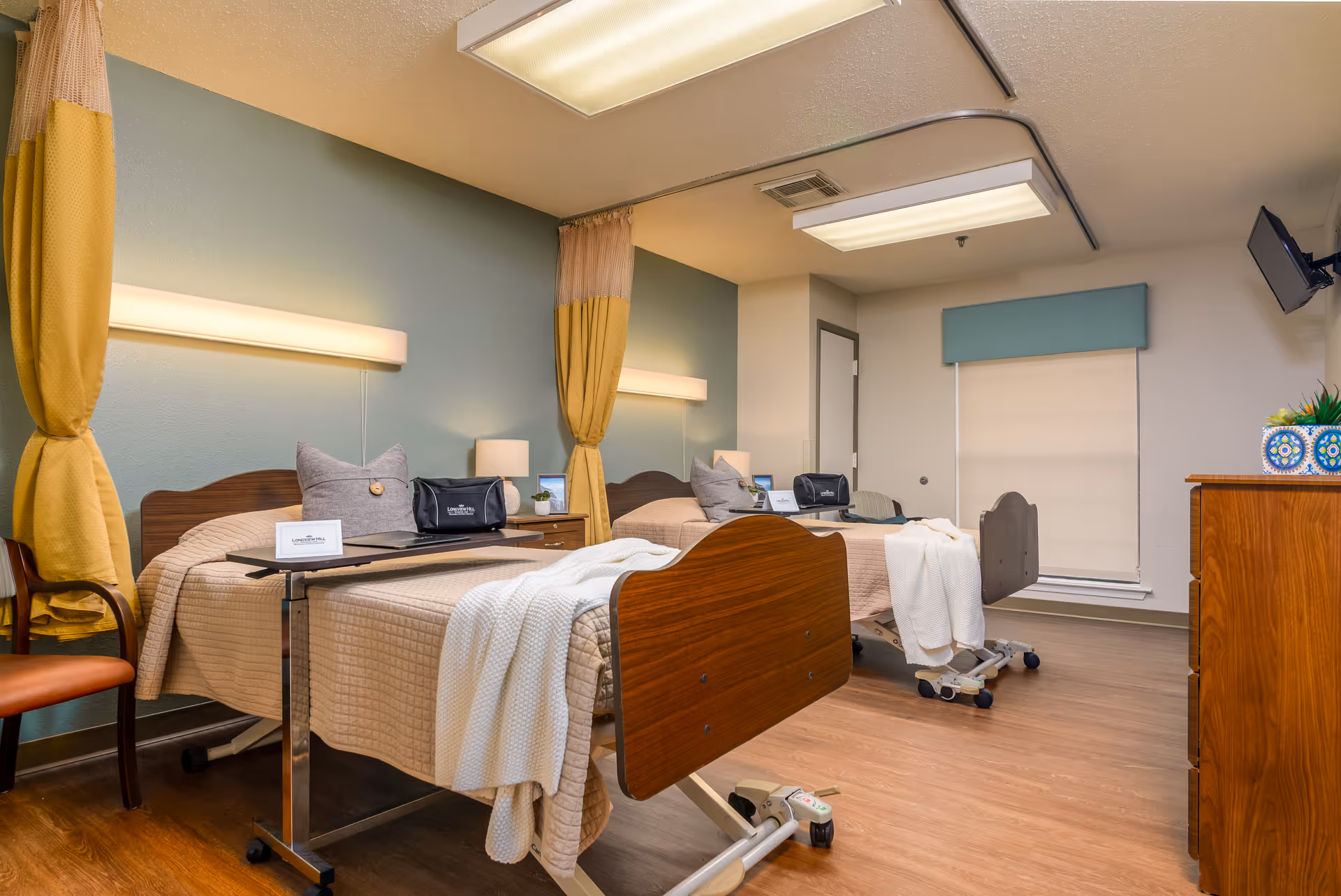 A bright nursing facility room with two adjustable hospital beds, bedside tables, privacy curtains, and a wall-mounted TV.