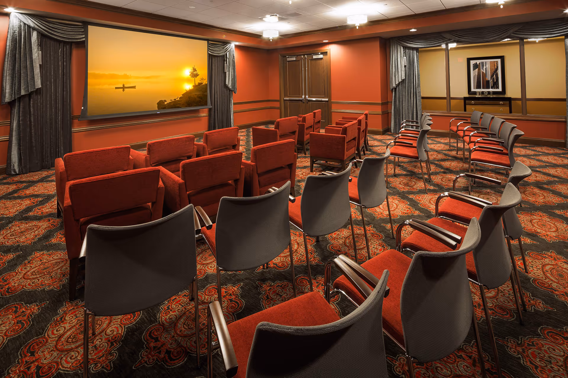 A small theater or presentation room with rows of red and gray chairs facing a large screen displaying a sunset scene with a person in a canoe on the water. The room has patterned carpet, red walls with wood trim, and gray curtains framing the screen and windows.