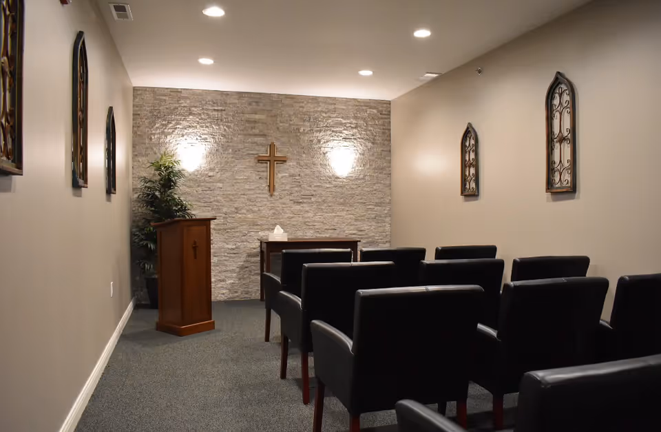 A small chapel or prayer room with rows of black chairs facing a wooden podium and a small table against a stone accent wall. A wooden cross is mounted on the wall above the table, and decorative wall hangings are on the side walls. The room is softly lit with recessed ceiling lights and wall sconces.