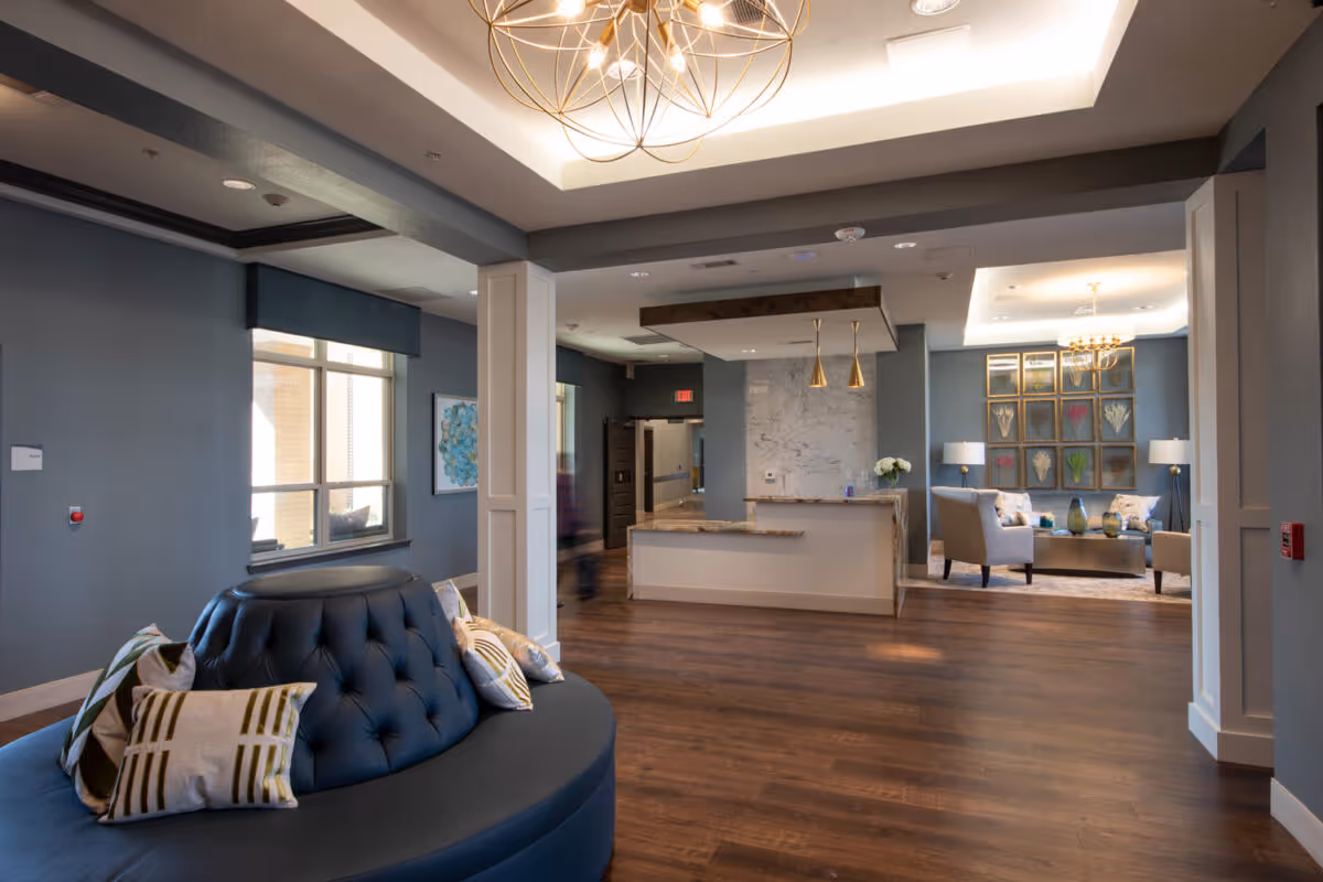 A spacious and modern common area in a senior living facility featuring a circular blue cushioned bench with decorative pillows in the foreground. The room has wooden flooring, blue-gray walls, and a reception desk with marble accents and gold pendant lights. In the background, there is a seating area with armchairs, a coffee table, lamps, and framed floral artwork on the wall. The ceiling has recessed lighting and a decorative chandelier.