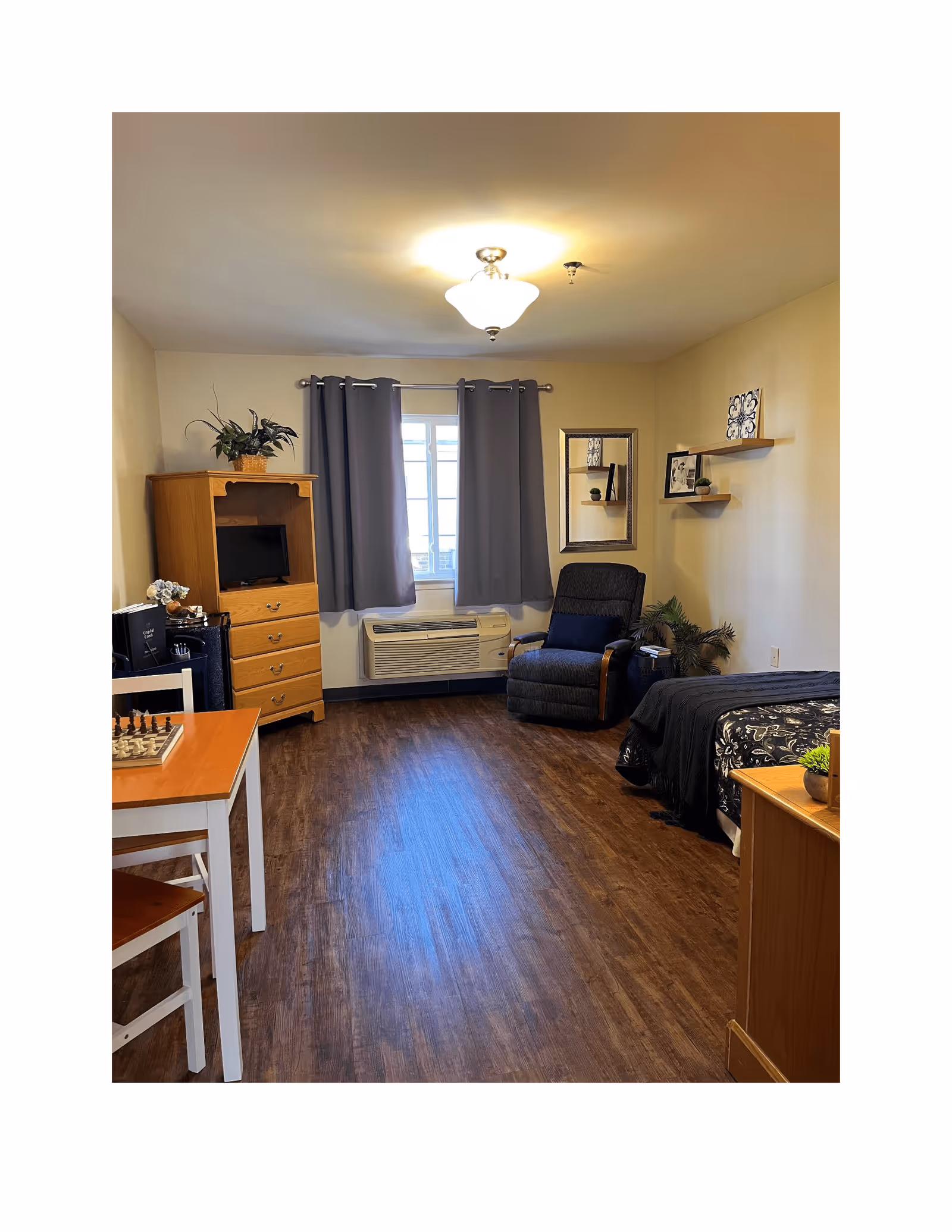 A cozy assisted living bedroom with wood flooring, a bed with black and white bedding, a cushioned armchair with a blue pillow, a wooden dresser with a small TV, a window with gray curtains, a small table with chairs and a chessboard, and decorative shelves with framed pictures and plants on the wall.