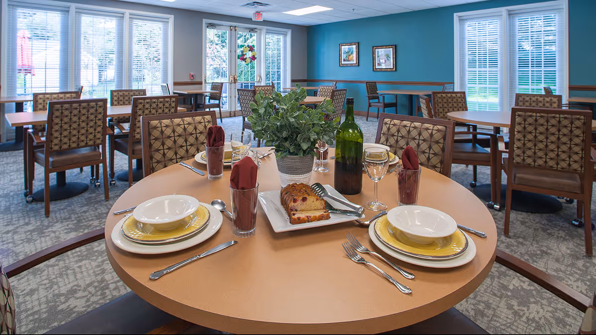 A dining room in a senior living facility with round and rectangular tables set with plates, bowls, glasses, and silverware. The round table in the foreground has a centerpiece with a green plant, a bottle of wine, and a plate with slices of cake. The room has large windows with blinds and blue walls with framed artwork.