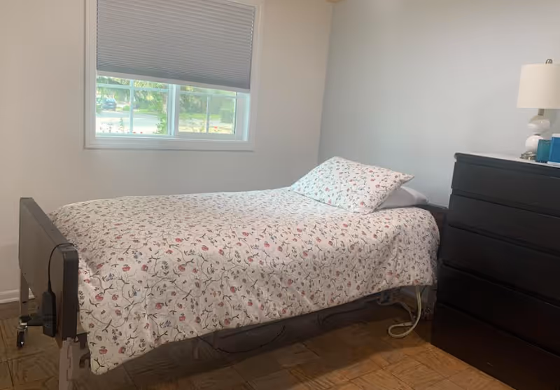 A small bedroom with a single bed covered in a floral patterned bedspread and a matching pillow. There is a window with a partially closed blind on the wall behind the bed. Next to the bed is a black dresser with a white lamp and some small items on top. The floor is wooden.