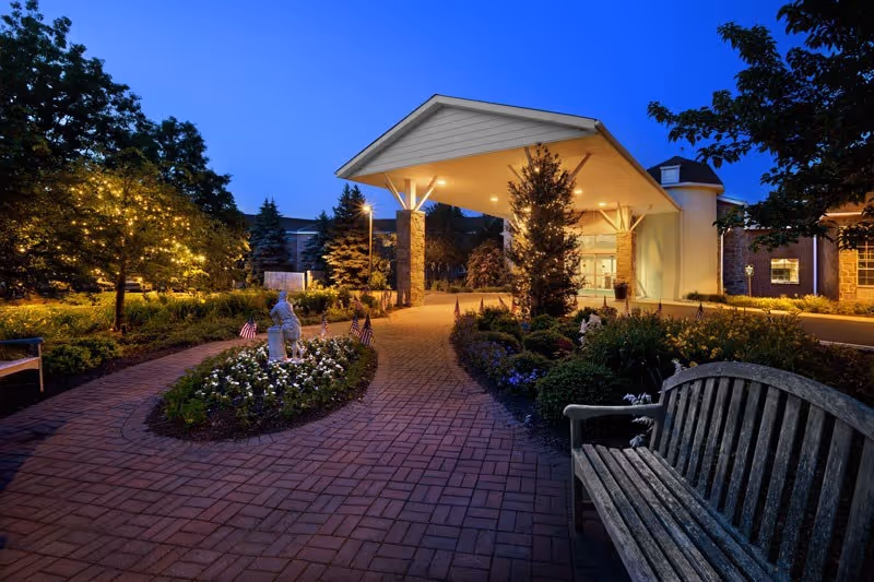 Evening view of the entrance to a senior living facility with a covered drop-off area, surrounded by landscaped gardens, small American flags, a statue, and benches along a brick pathway, illuminated by soft outdoor lighting.