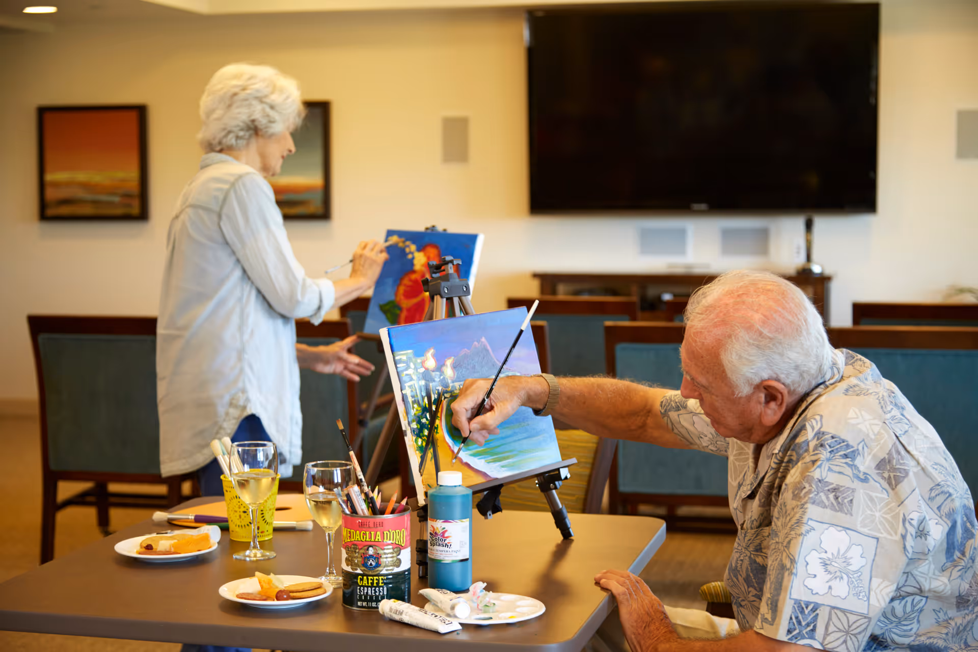 An elderly man and woman are painting on canvases set on easels in a well-lit room. The man is seated at a table with painting supplies, including brushes, paint tubes, and glasses of white wine, while the woman stands in the background working on her painting. The room has chairs, framed artwork on the walls, and a large flat-screen TV mounted on the wall.