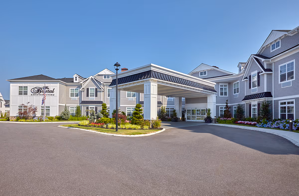 Porte-cochère and landscaped circular driveway at the front of a multi-story assisted living building under a clear blue sky.