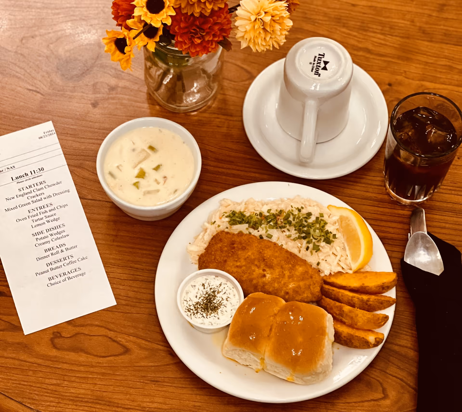A wooden table set with a meal including a plate of fried fish, rice garnished with herbs, potato wedges, two dinner rolls, and a lemon wedge. There is a small cup of tartar sauce on the plate. Next to the plate is a bowl of New England clam chowder, a glass of iced tea, and an upside-down white coffee cup on a saucer. A printed lunch menu and a small vase with orange and yellow flowers are also on the table.